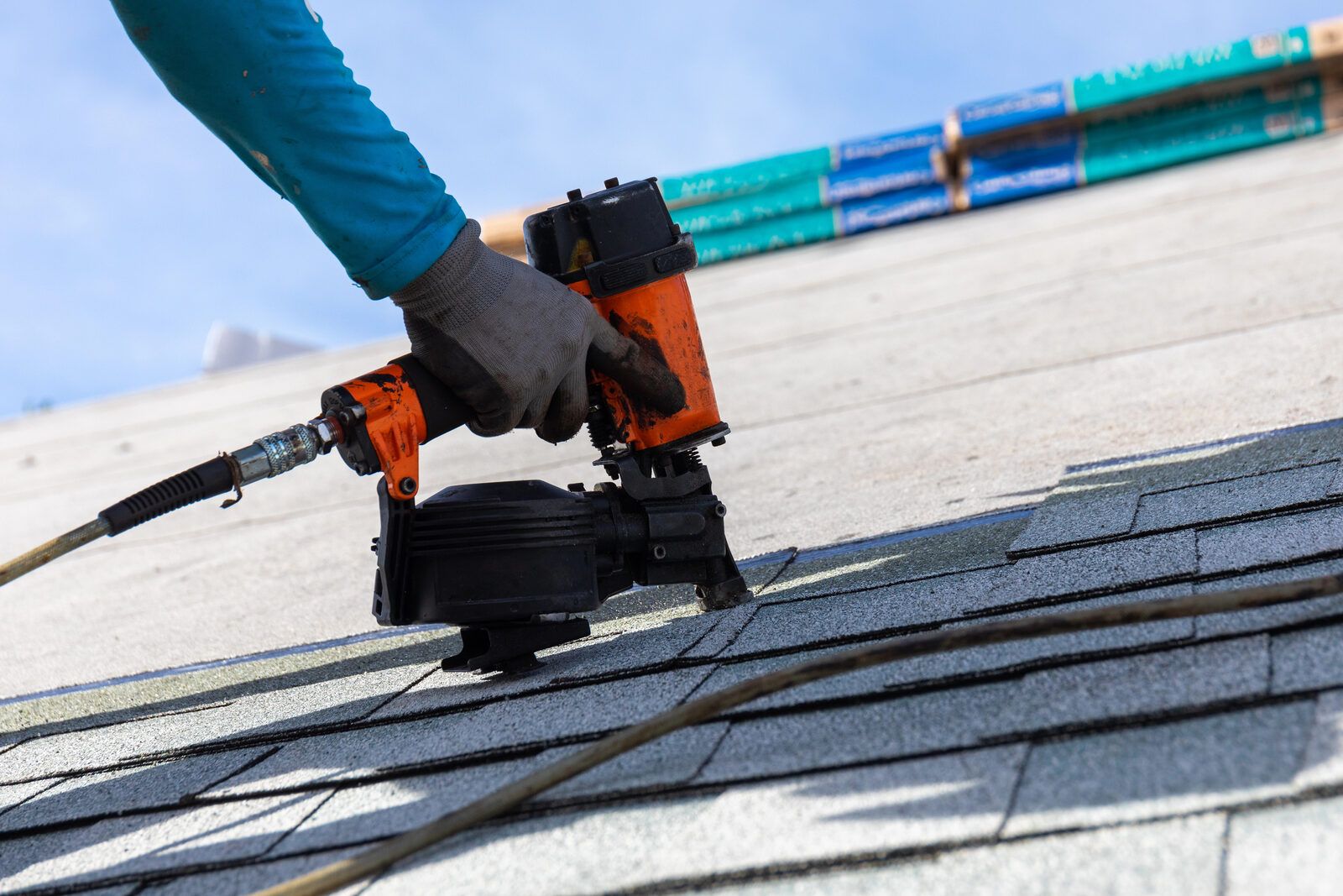 A gloved hand holding a coil roofing nailer over a fresh course of grey asphalt shingles.