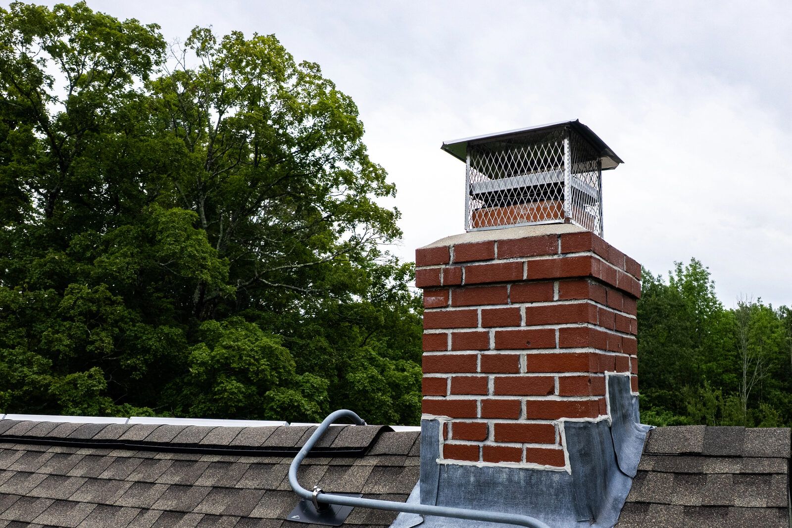 A brick chimney with metal flashing and a cap on a shingle roof.