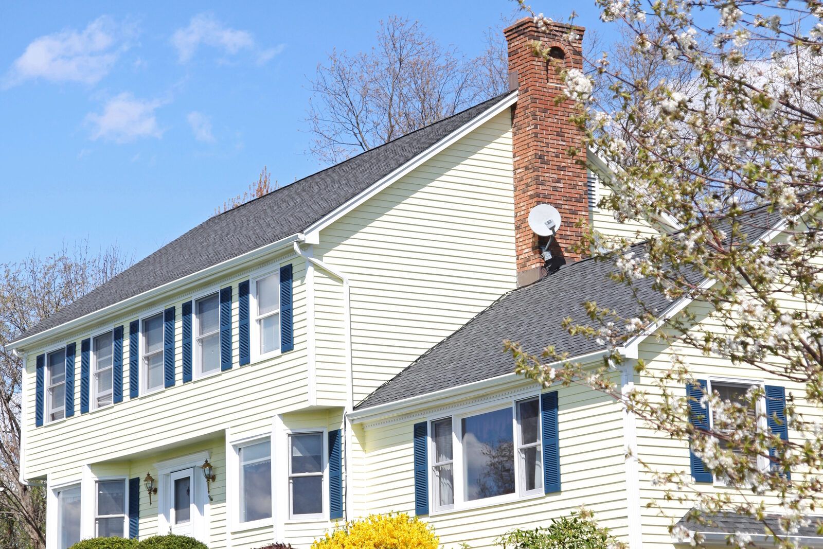 A two-story clapboard house with a dark grey shingle roof and a brick chimney in spring light.