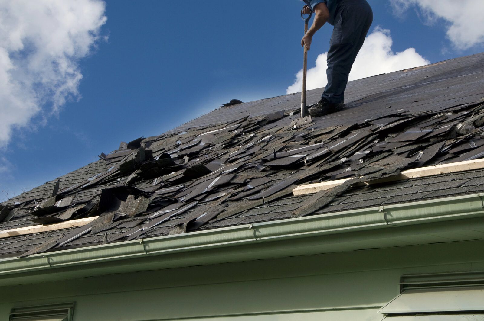 A roofer stripping old shingles from a roof, with torn shingles piled along the eave.